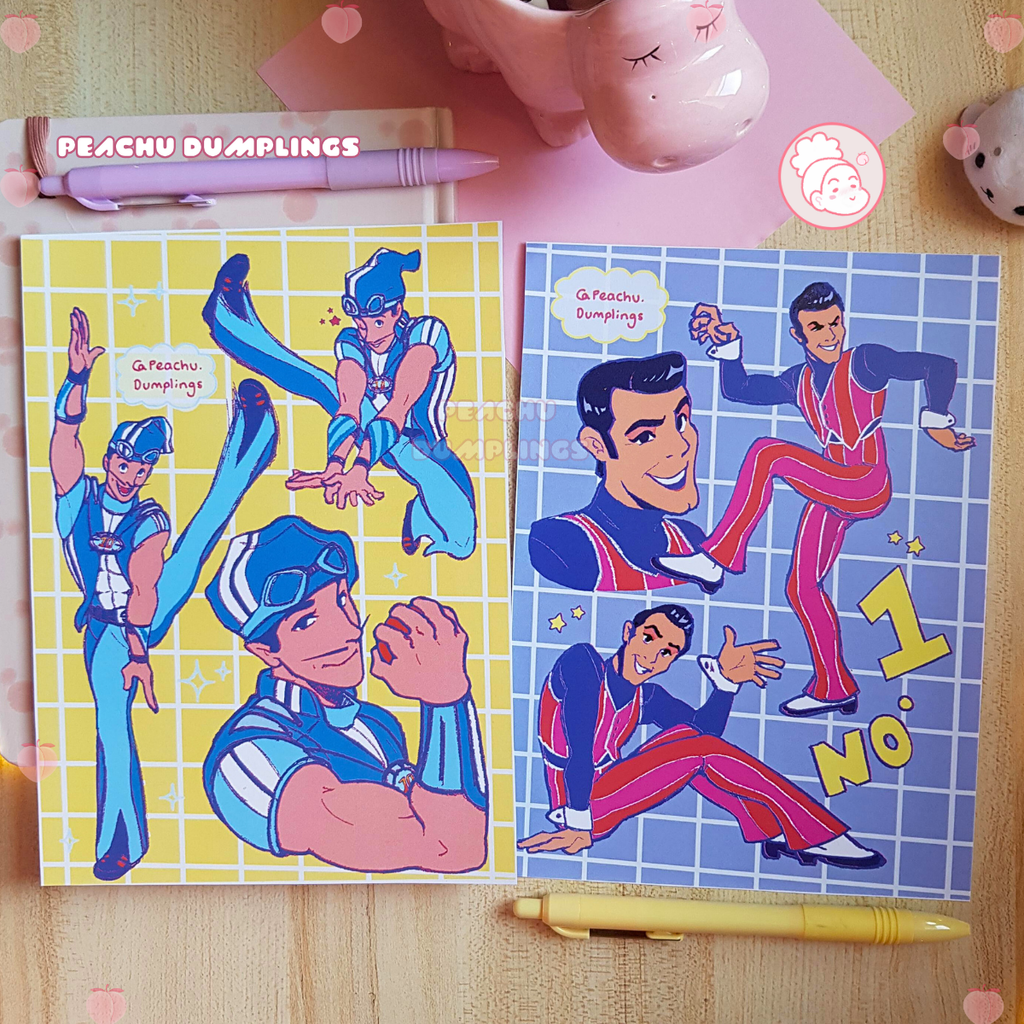 Two illustrated cards featuring characters in colorful outfits on a wooden surface with pens and a small figurine.